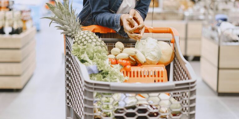 A shopper's hands resting on a cart full of fruit and vegetables in a supermarket aisle