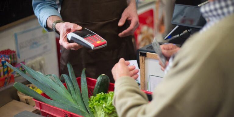 A shopper pays by card at a grocery checkout, with leeks, lettuce and a cucumber in a red basket